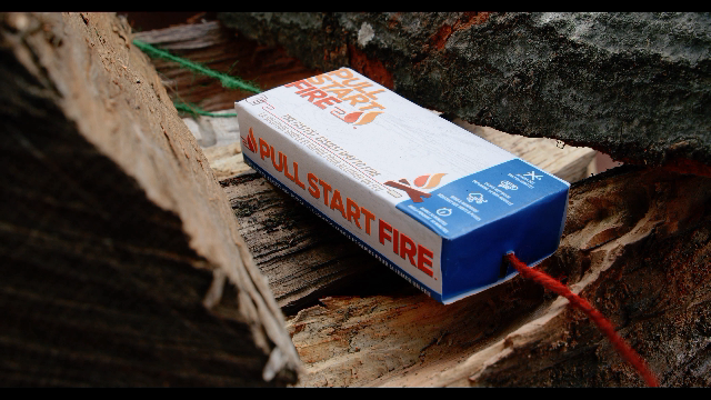 
                  
                    Pull Start Fire firestarter placed between firewood logs, showing red pull string for easy ignition
                  
                